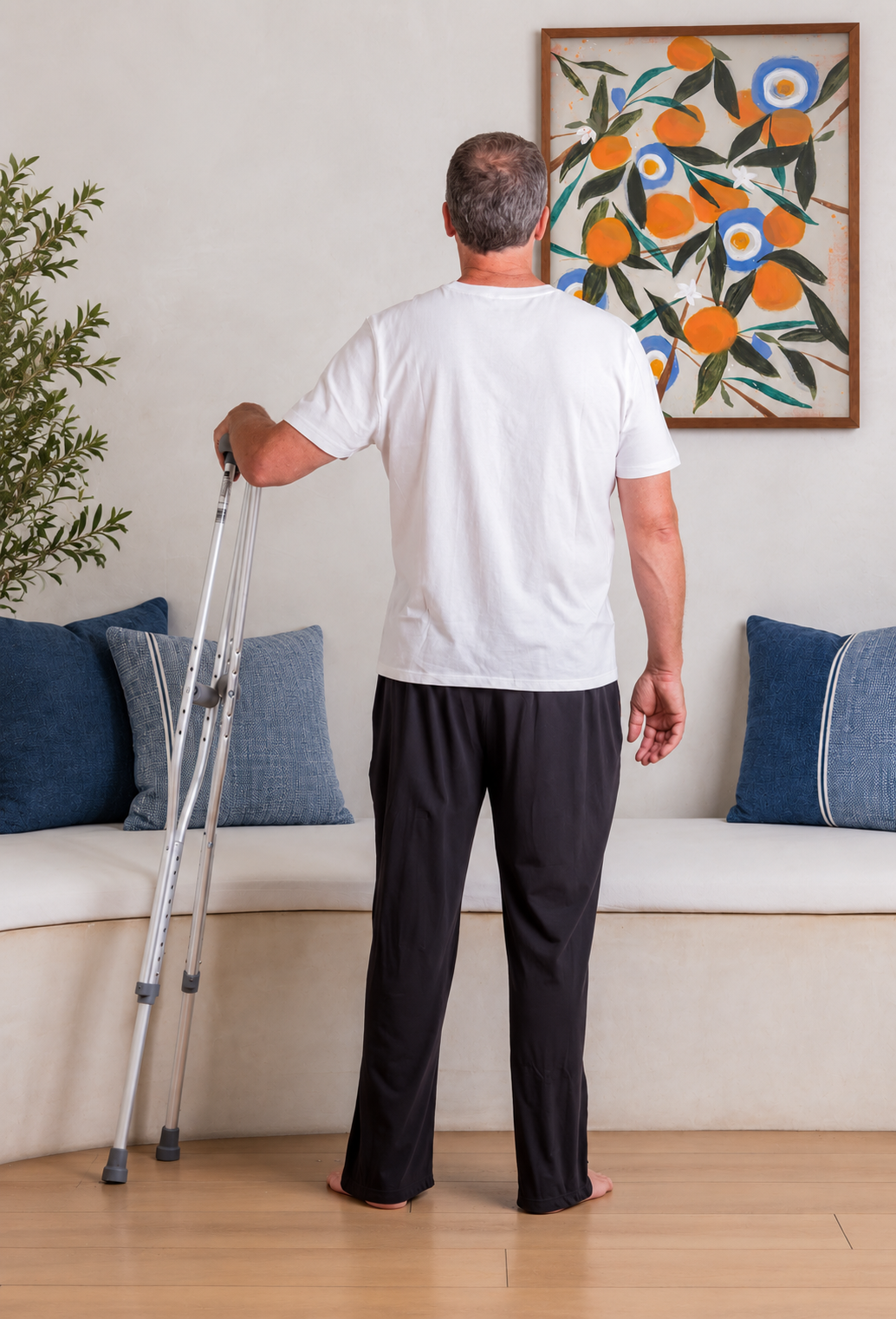 A man viewed from behind stands in a living room wearing a white t-shirt and black adaptive tearaway pants, holding a pair of crutches next to a beige sofa with blue pillows.