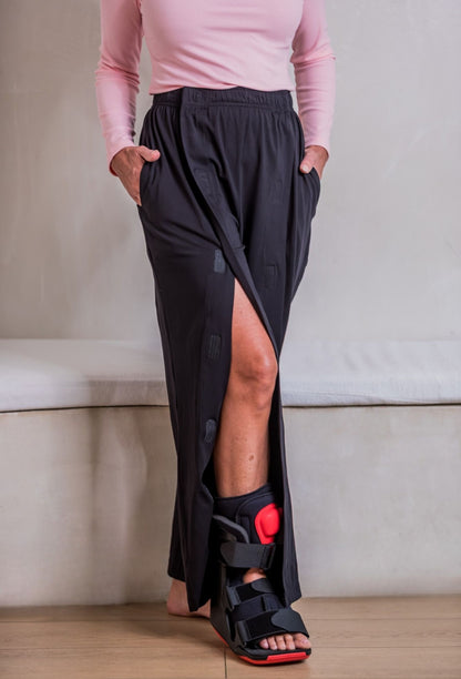 A person wearing loose black adaptive velcro pants and a black ankle support brace, paired with a light pink long-sleeve top to demonstrate how the pants can be worn to work or errands, standing indoors against a light neutral wall.