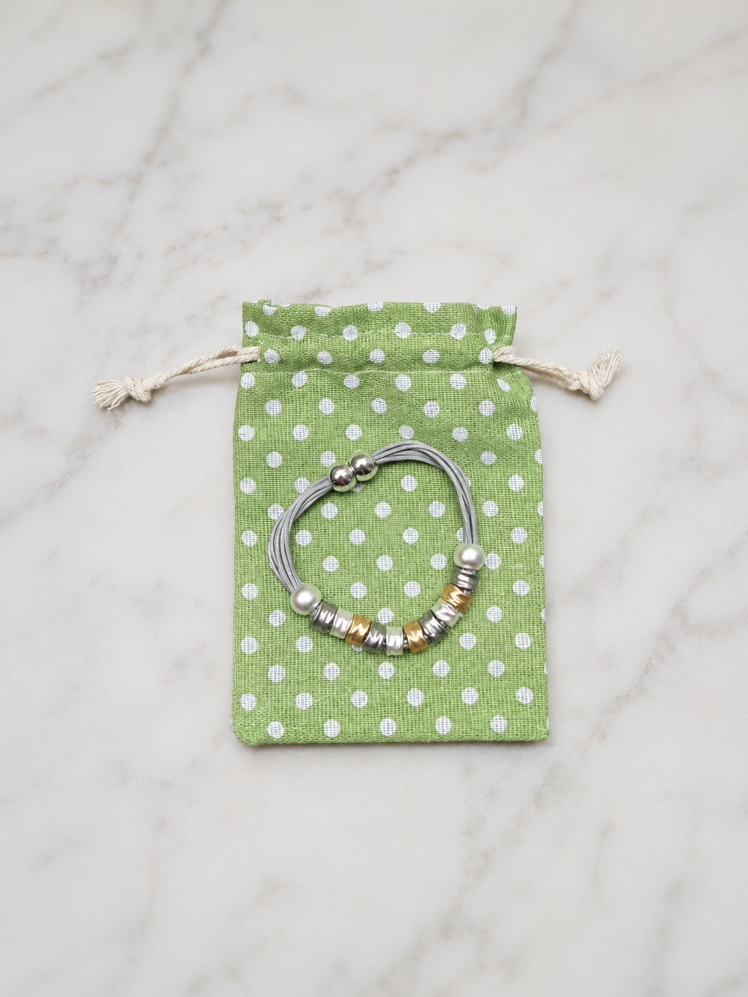 A silver-toned beaded bracelet with white and gold beads, displayed on a green and white polka dot drawstring pouch over a white marble surface.