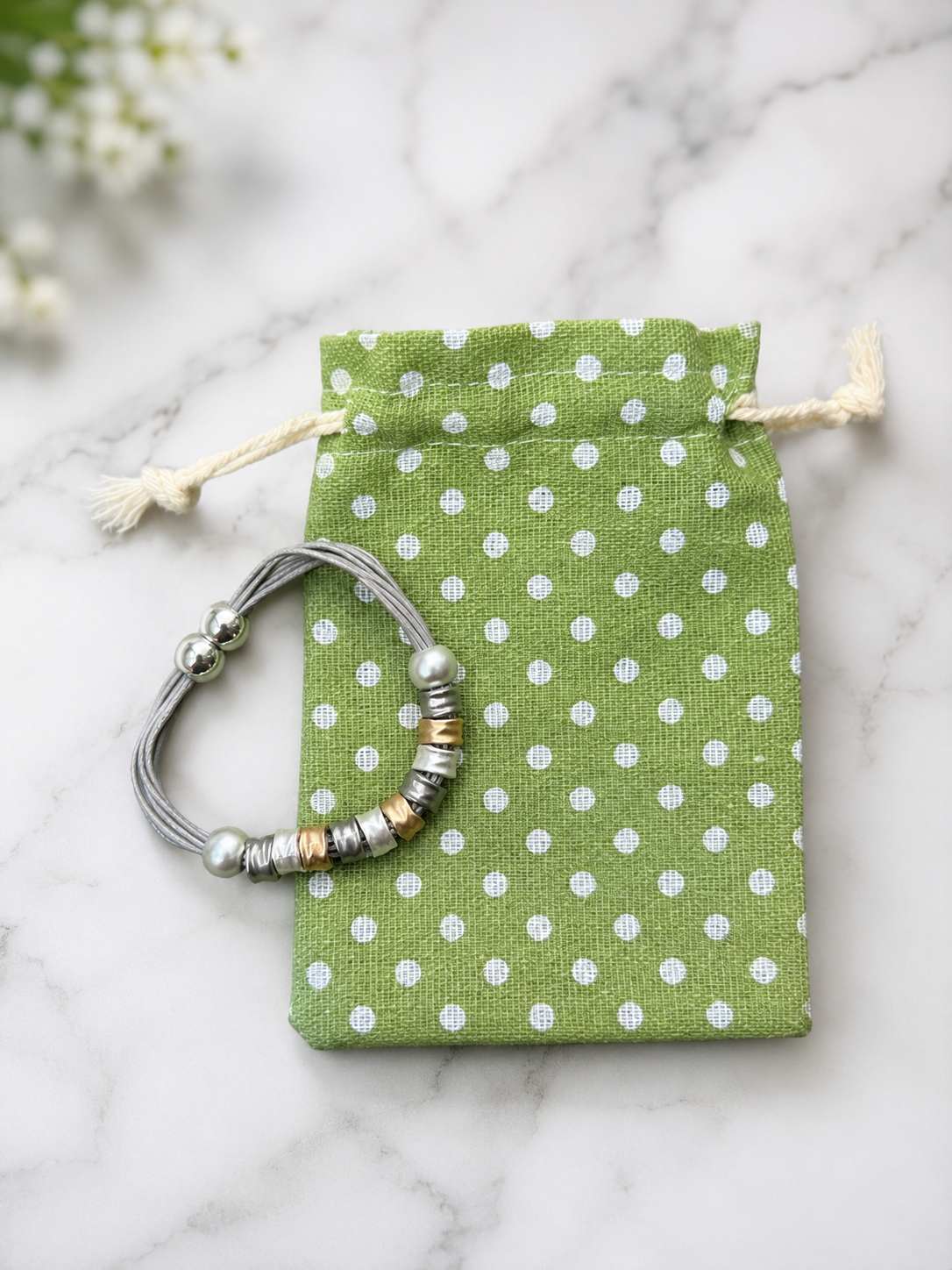 A silver beaded bracelet with metallic accents is displayed on a small green drawstring pouch with white polka dots, set on a light marble surface.