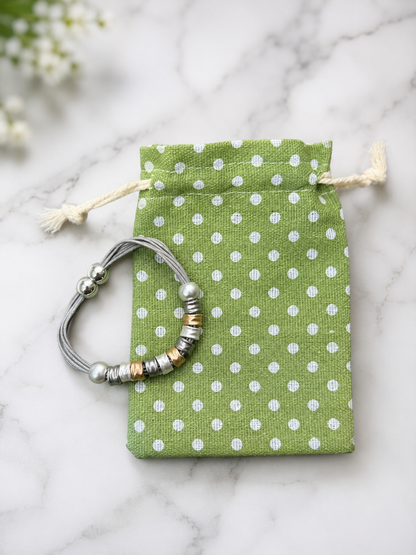 A silver beaded bracelet with metallic accents is displayed on a small green drawstring pouch with white polka dots, set on a light marble surface.