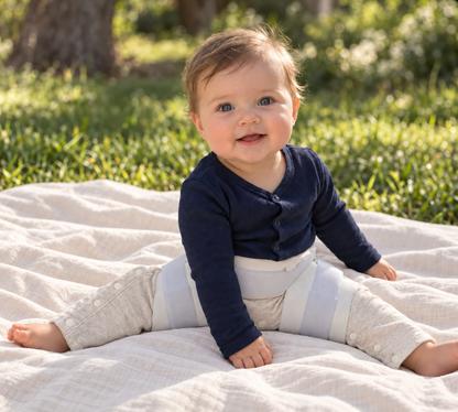 A baby sitting outdoors on a light beige blanket, wearing a navy blue long-sleeve henley top and light beige hip dysplasia pants and constructed Pavlik harness over the pants