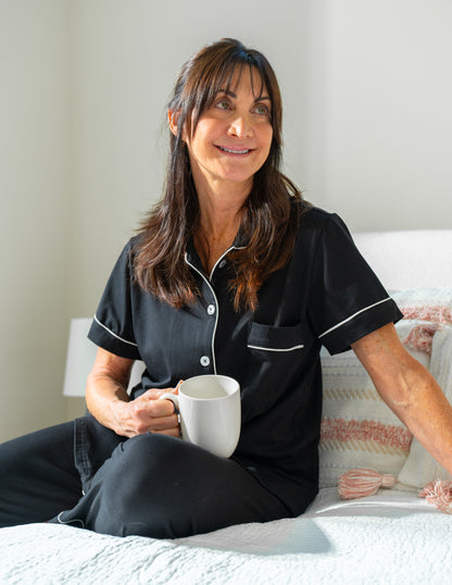 Woman in black surgery recovery pajamas holding a white mug on a bed.