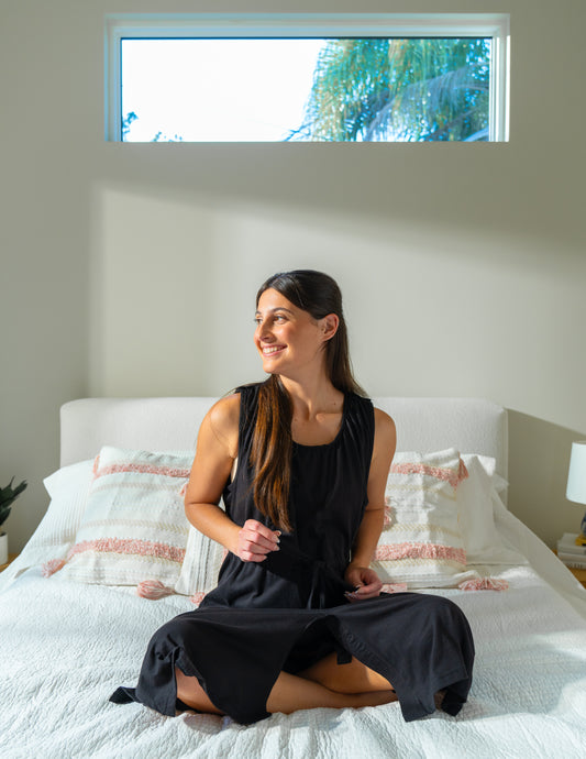 Woman sitting on a bed wearing the black hospital gown showing the string which makes it an adjustable waist