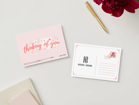 A flat-lay of two postcards on a light gray surface. One postcard is light pink with the words 'thinking of you' in rose-gold script and small heart accents.