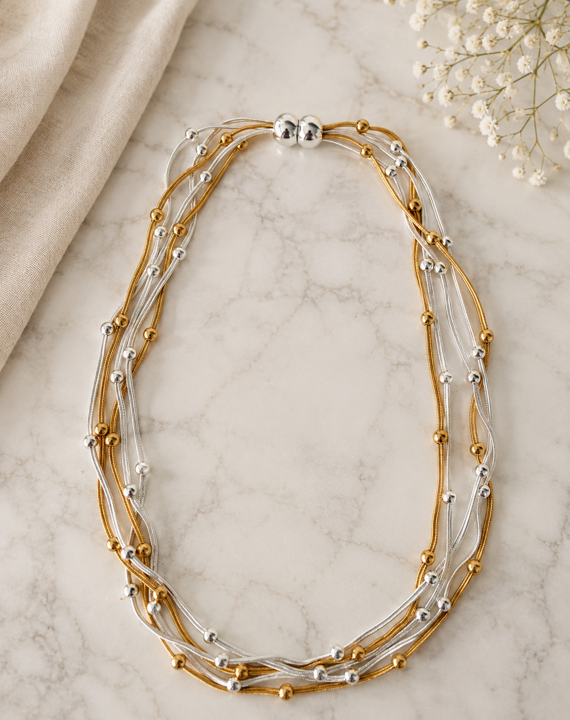 A multi-strand necklace with gold and silver-tone cords, accented with small metallic beads, displayed on a light marble surface with a beige fabric and white flowers in the background.