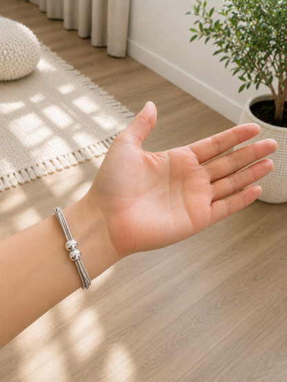 A silver-toned metal bracelet with two small round beads worn on a person's wrist, shown in a bright indoor room with wood flooring, a potted plant, and a woven rug.