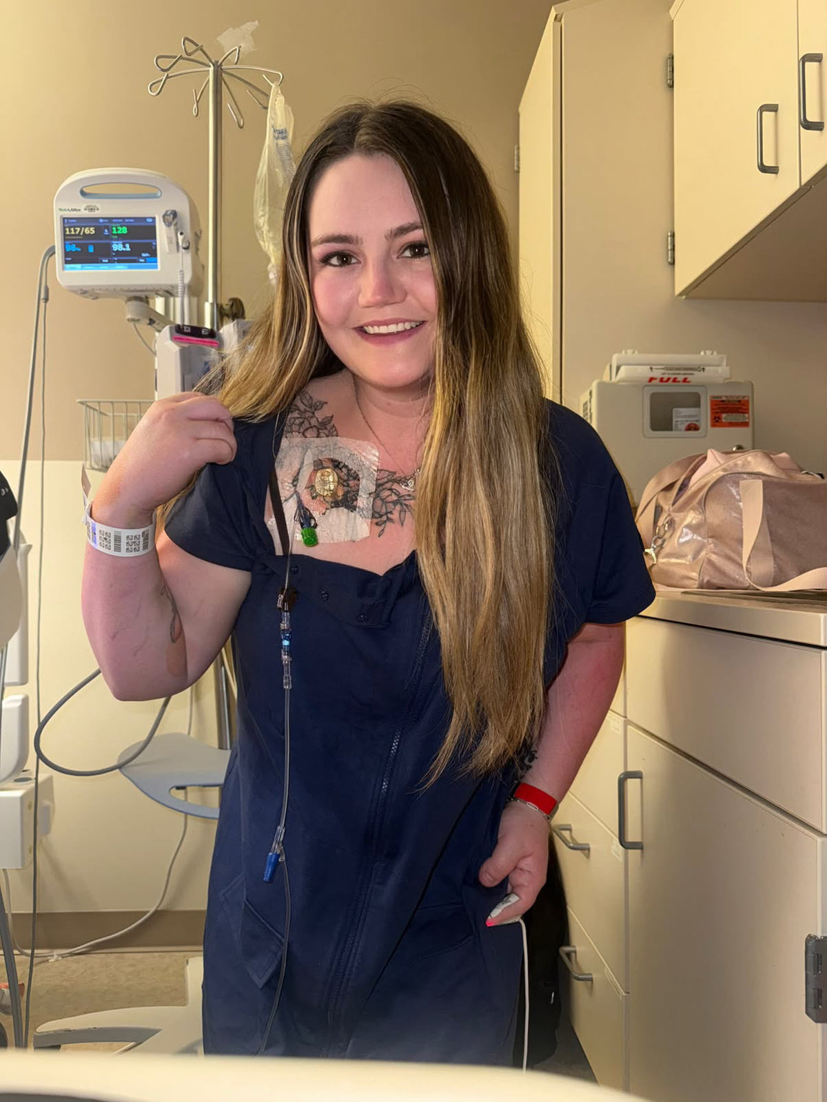 A woman with long blonde-highlighted hair stands in a medical room wearing a navy blue hospital gown, showing the chest panel open and her port for chemotherapy. Medical equipment, cabinets, and an IV stand are visible in the background.