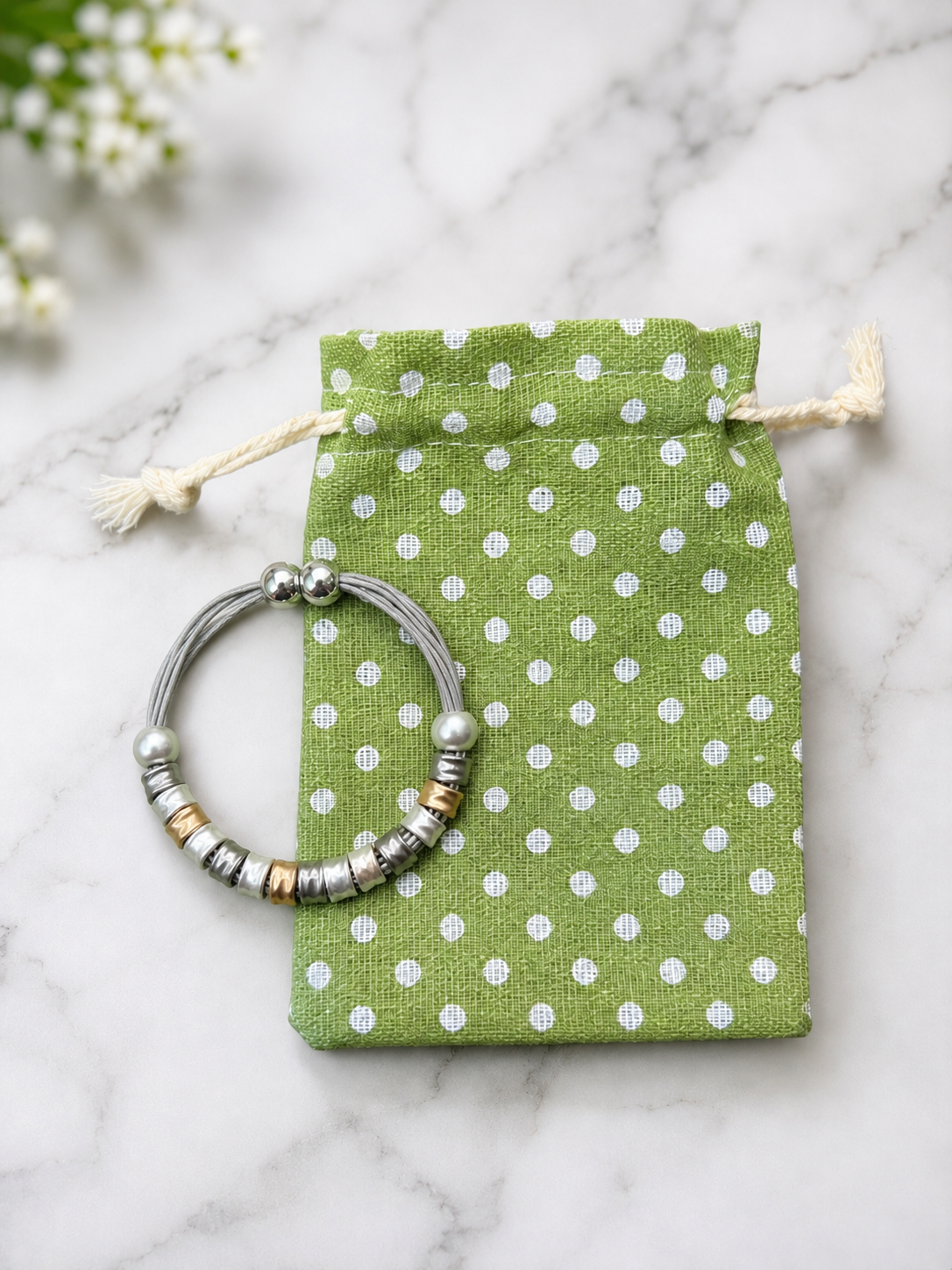 A silver-toned beaded bracelet with metallic spacer beads rests on a small green drawstring pouch with white polka dots, placed on a light marble surface with blurred white flowers in the corner.