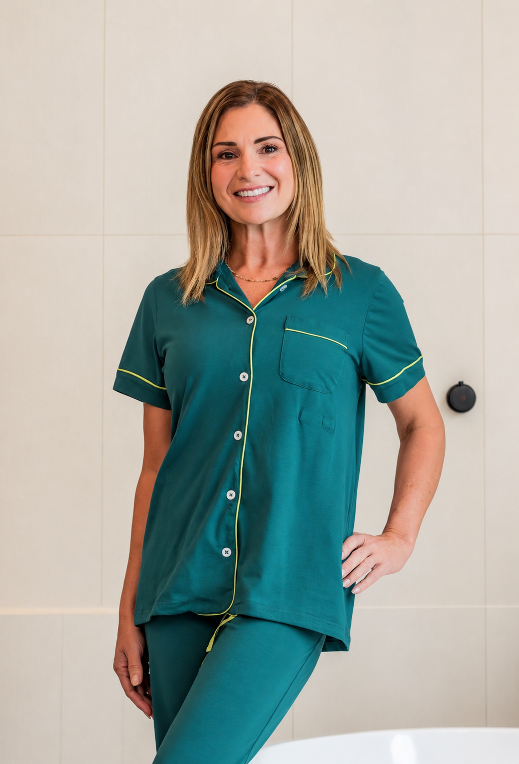 A woman wearing a teal short-sleeve pajama shirt with green piping and matching teal pajama pants, standing against a light beige tiled wall.
