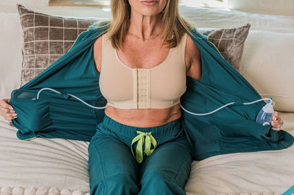 A woman sits on a bed teal surgery recovery pajamas, holding both sides of the top open to reveal surgery drain bulbs in the pockets.