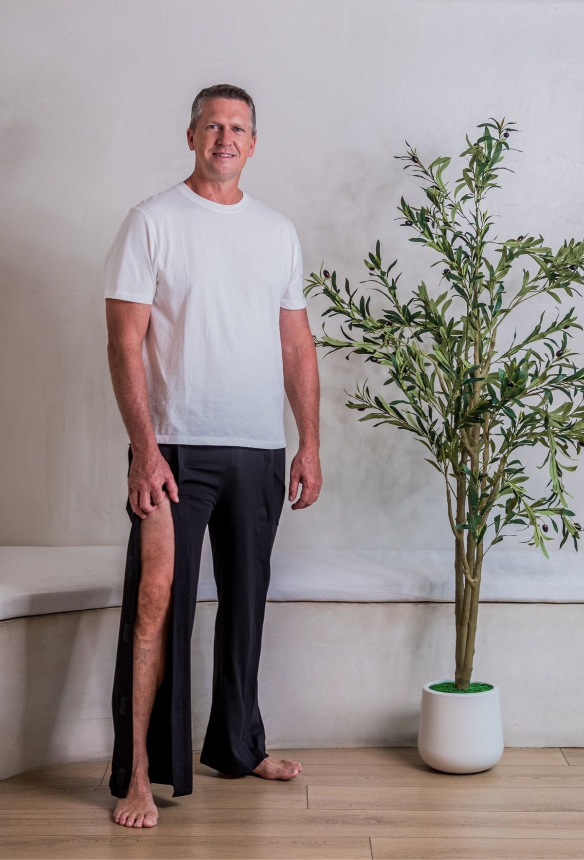 A man wearing a white crewneck t-shirt and black tearaway pants, open up high to show leg accessibility for things like physical therapy, suture or stitch removal, braces and immobilizers, and leg IV access.