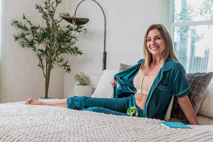 A woman sits on a bed in a bright bedroom, wearing a teal short-sleeve pajama top and matching pants over a beige bralette. She is holding open the pajama top, to show internal pockets which hold a surgical drain bulb and clipped in tubing.